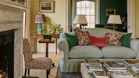 Elegantly styled living room featuring a light blue skirted sofa with colorful accent pillows, a marble fireplace, an upholstered coffee table stacked with books, and an doorway opening into a teal room.