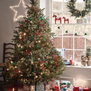 Traditionally decorated Christmas tree next to window, red and green decorations, stairs, wreath, wooden horses, presents under tree