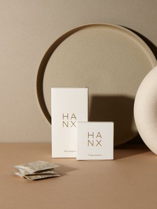 Neutral tone background, base and circular tray on far wall, two small neutral boxes with HNAX logo, four stacks condom sachets left of the boxes