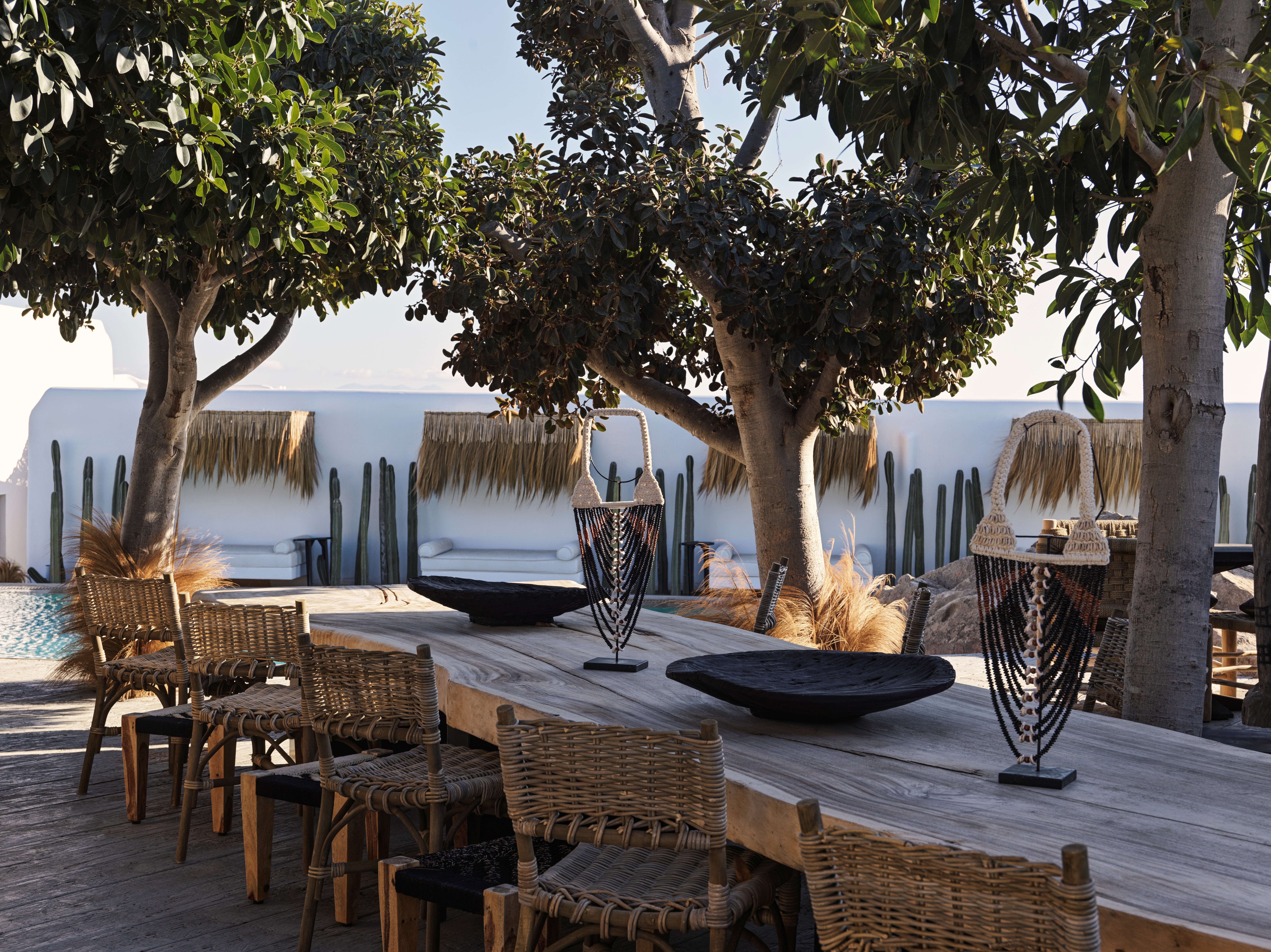 A long wooden table placed in a beach club, shaded by trees baking in the sun, features a series of rattan and wooden furniture pieces, central organic black bowls, and rustic accessories.