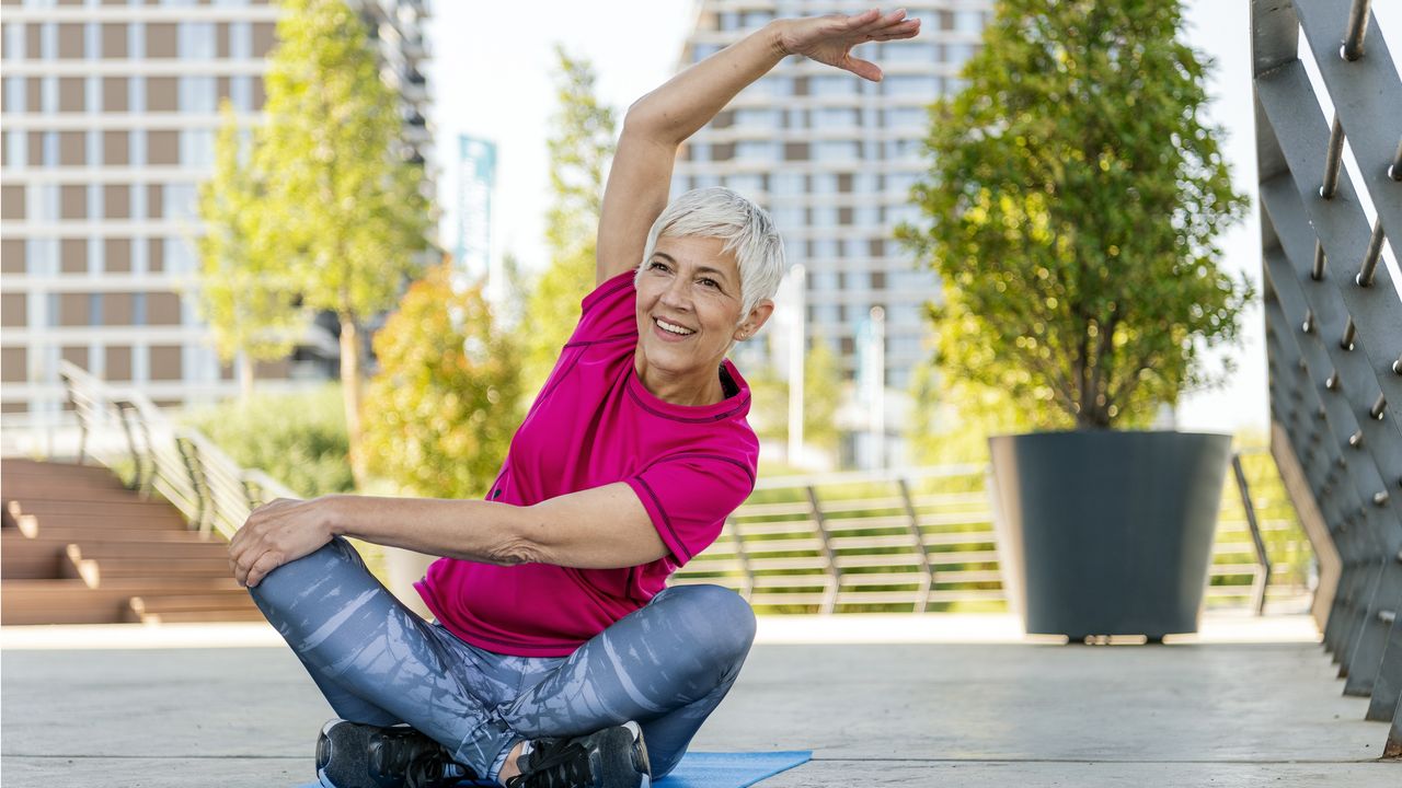 An older woman sits outside on a mat doing yoga.