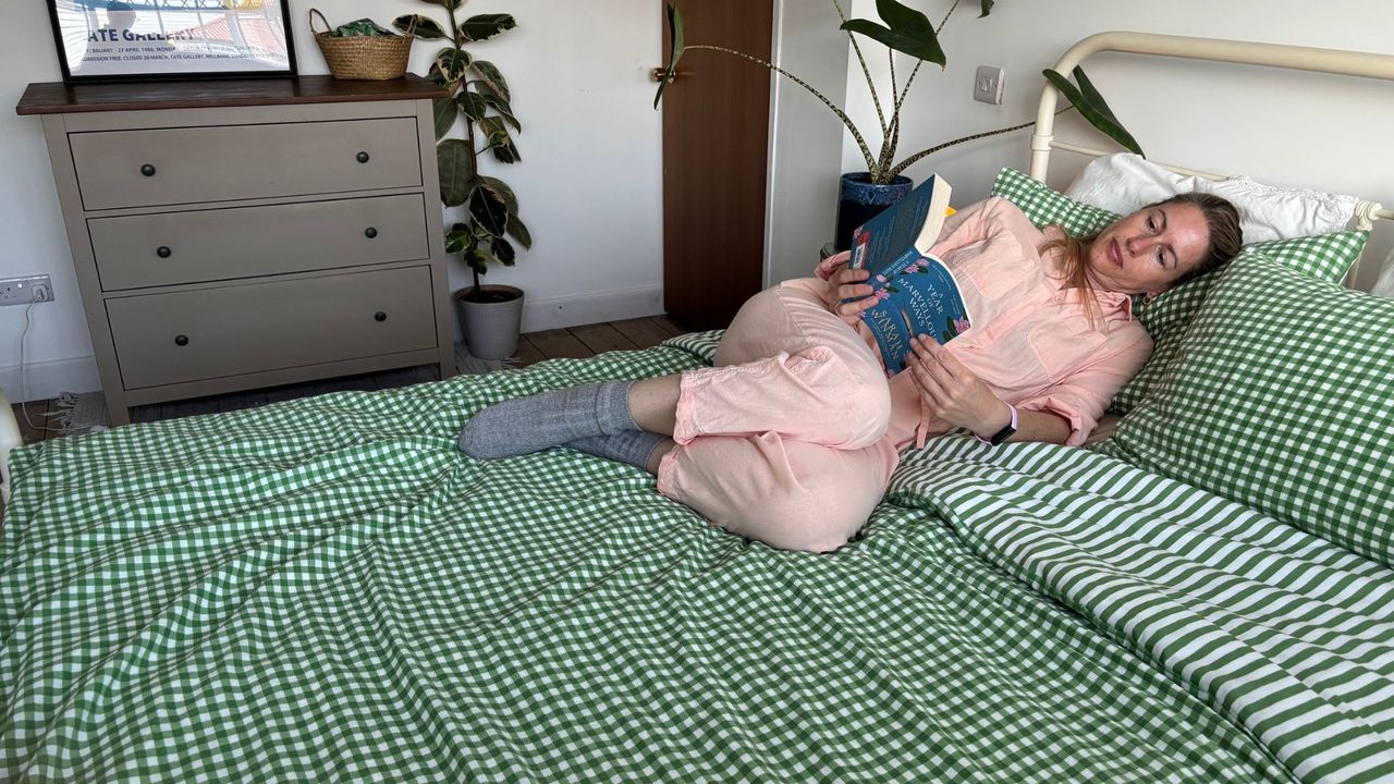 The image shows a mattress tester lying on the Birch Elite Natural mattress, which is dressed in green and white gingham bedding, while reading a book