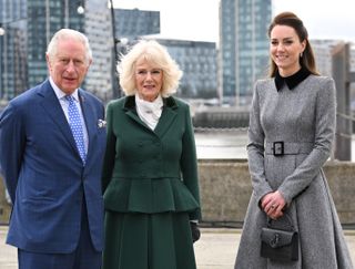 Kate Middleton wearing a gray dress standing next to The King and Queen along the river with buildings behind them