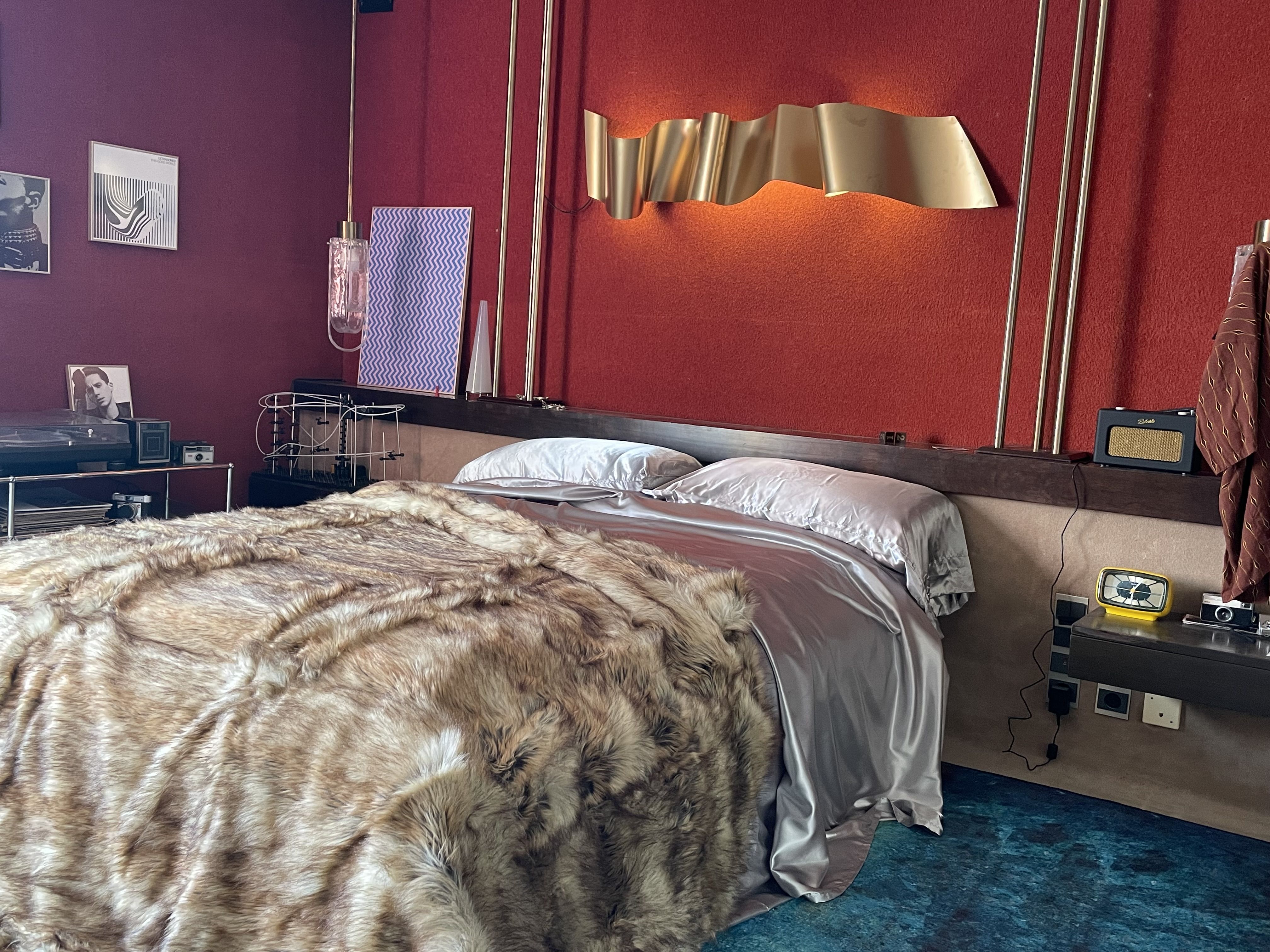 A sumptuous bedroom designed with a red-painted wall, a brown one, lots of golden decor, leopard-print bedsheets, scattered objects, and a teal rug.