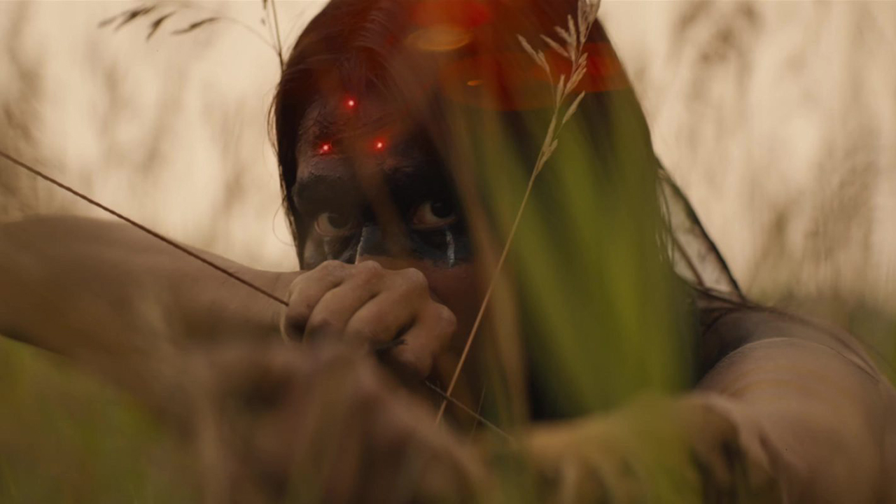 Close up of a woman with black war paint over her eyes. She is pulling back a bow. On her forehead there are 3 red dots from the Predator's sniper sight.