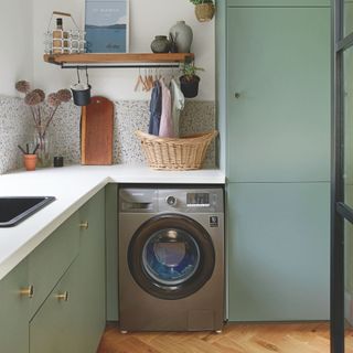 Kitchen with green cabinets and a grey washing machine , with white worktops and terrazzo-style backsplash on the wall