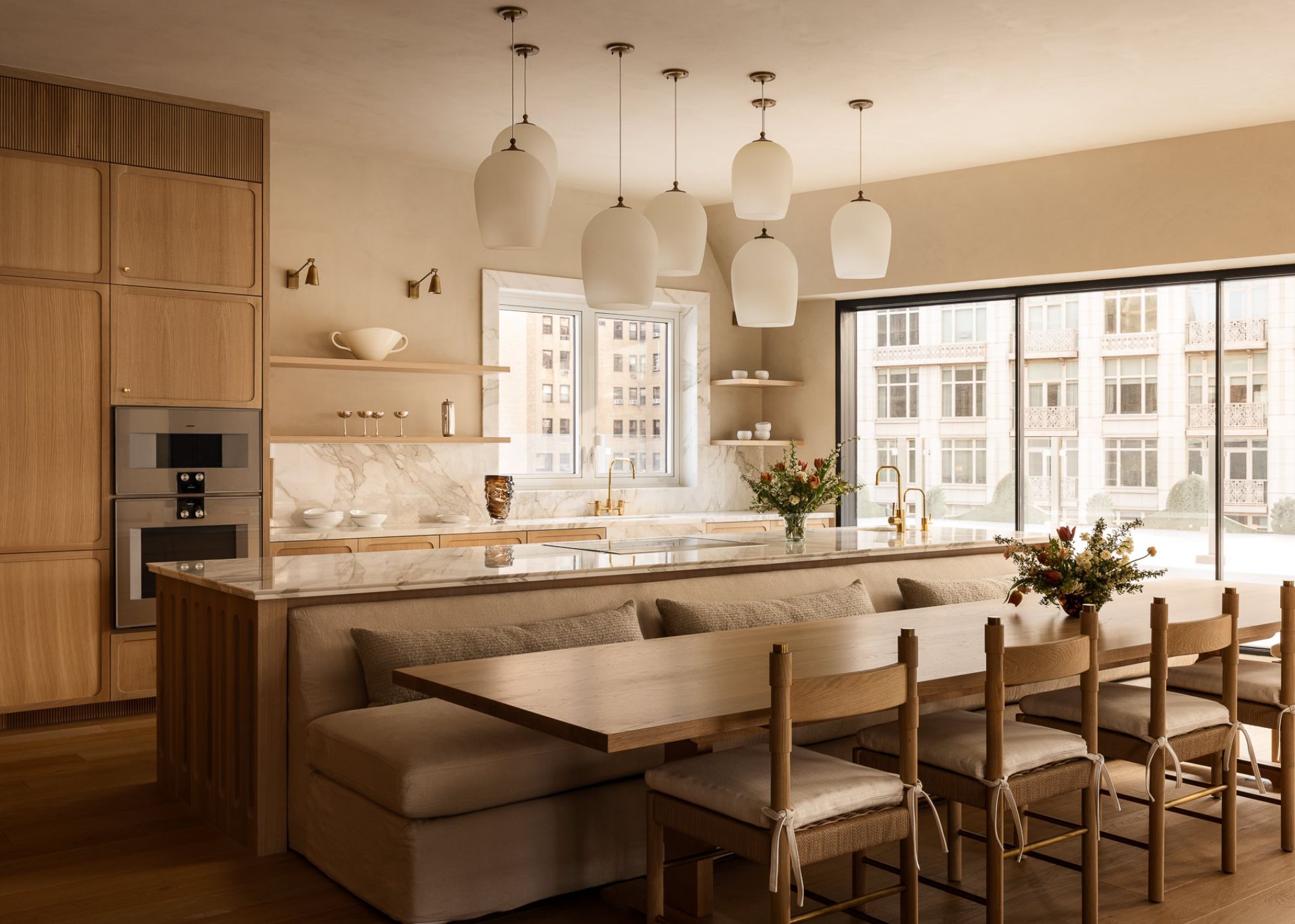 A modern timber kitchen with fluted detailing on the cabinetry, a marble worktop and island with several pendant lights hanging above and with built-in seating on the dining table side