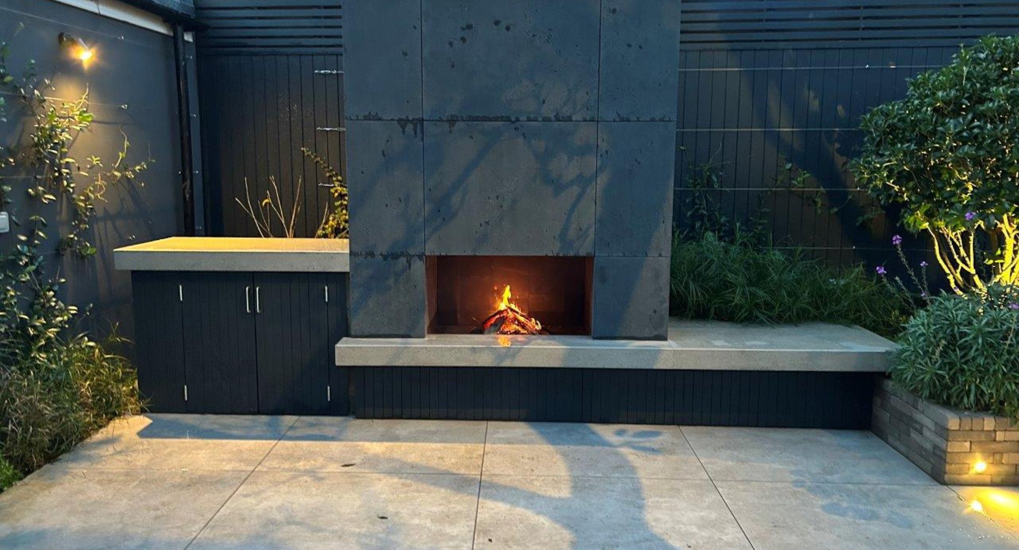 Urban patio garden with a grey fireplace and shrubs surrounding it. LED lights also cast shadows onto the tiles