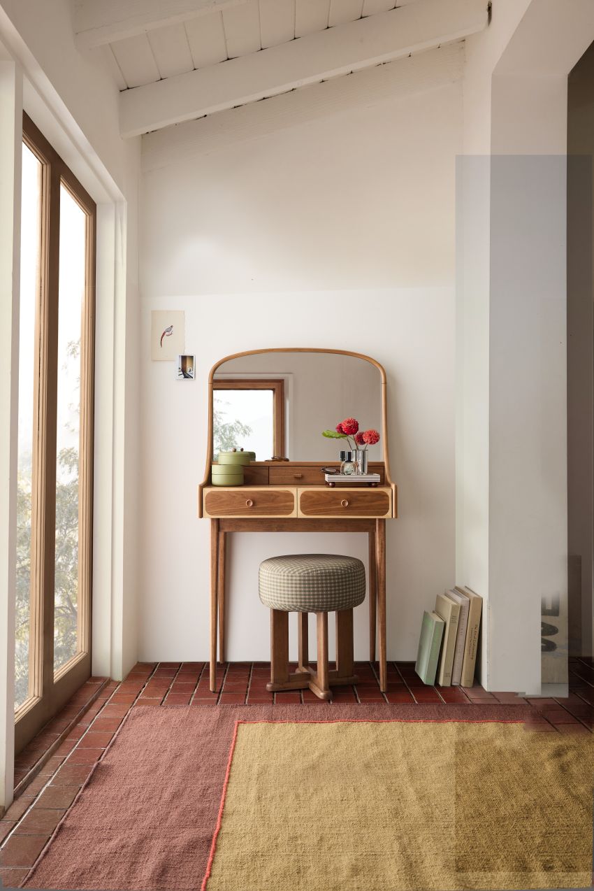 Image of a wooden mid-century-inspired vanity set in a white room with large windows on the left wall and brick flooring that has a green and terracotta rug on it. 