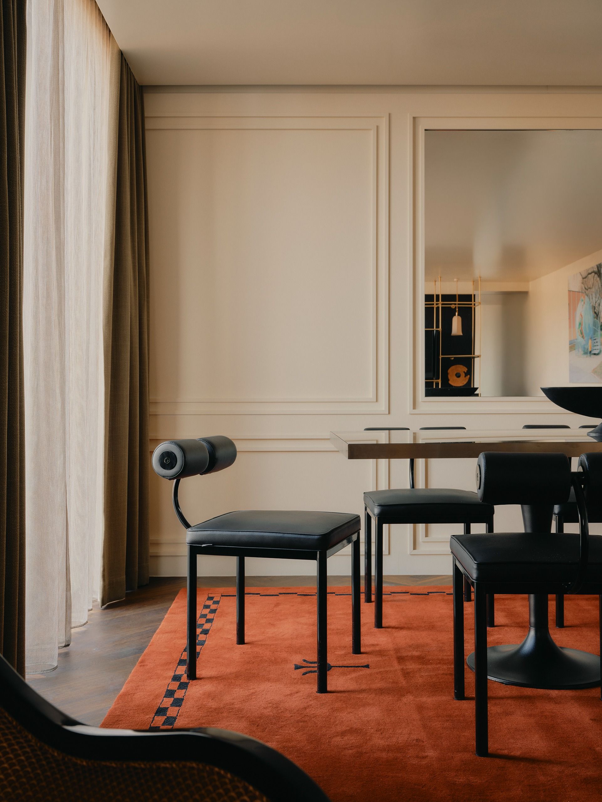 A modern dining area with black roll-back dining chairs on top of an orange rug with a black checkerboard border in front of double-layered curtains with sheer underneath and green linen on top