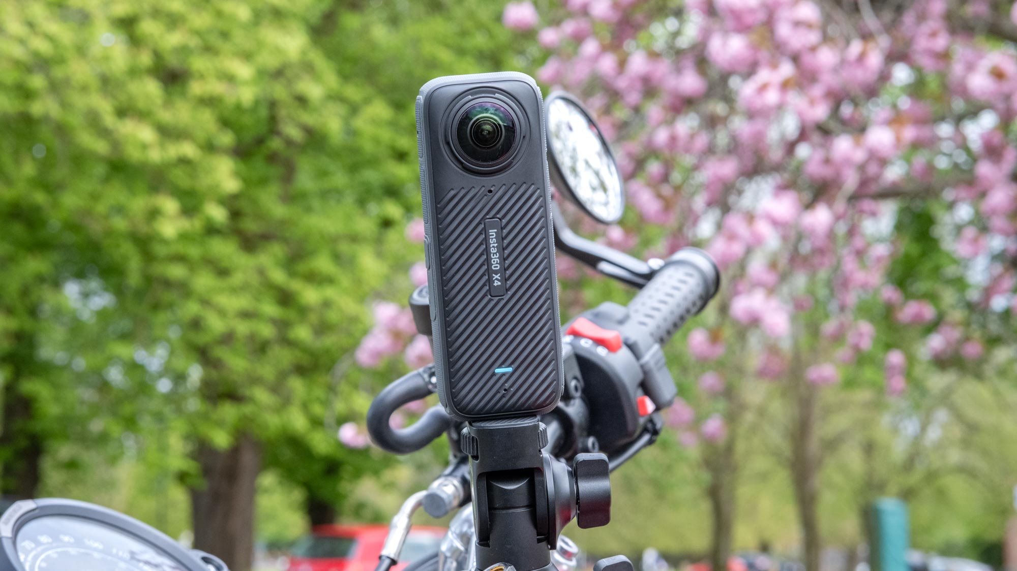 A photo of the Insta360 X4 mounted on motorcycle handlebars with pink and green foliage in the background.