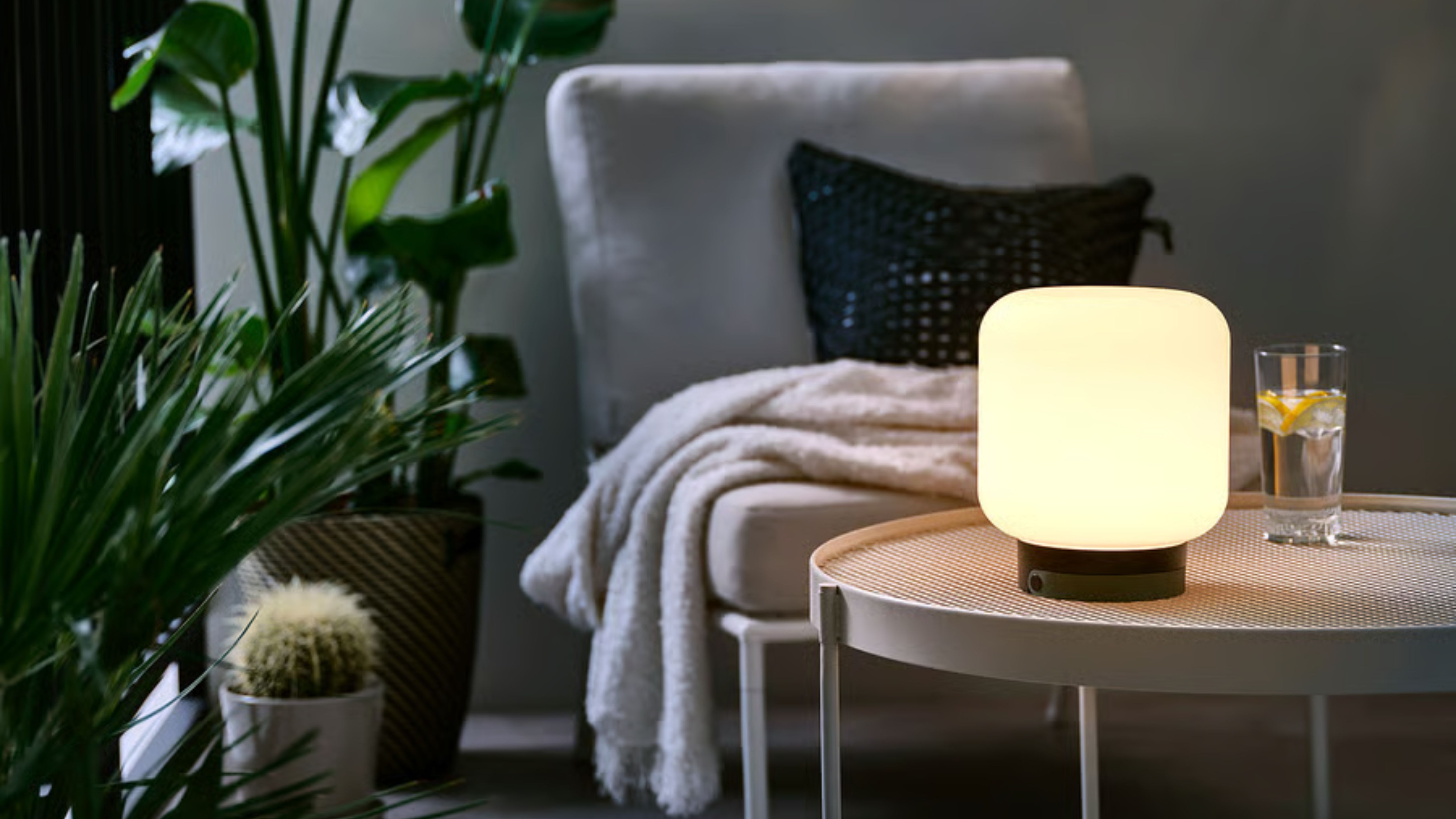 IKEA outdoor L&Auml;NSPORT lamp pictured atop of a wooden patio table alongside a lounge chair and various potted plants.