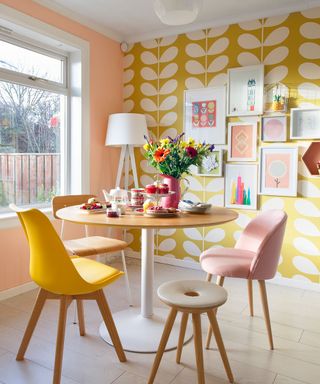 dining area with pink and yellow wall with pictures frame and white large window and wooden floor with round dining table with multicolour chairs