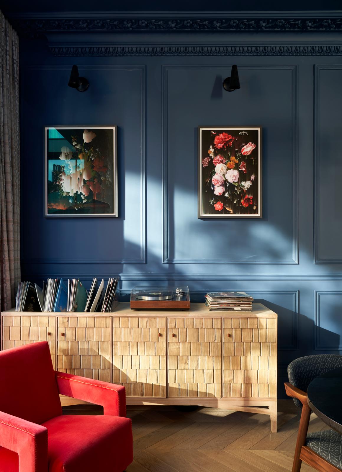 Image of a dark blue living room with a custom, wood vinyl storage cabinet. There are two art pieces hanging on the wall in crown molding with a museum light above each of them. A red accent chair is in view.