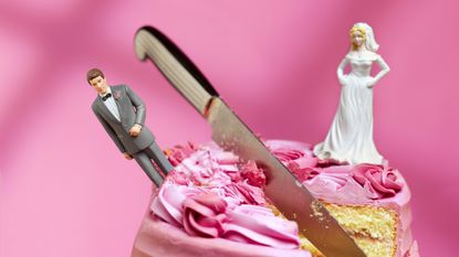 A knife cuts through a pink wedding cake between the bride and groom figurines on top.