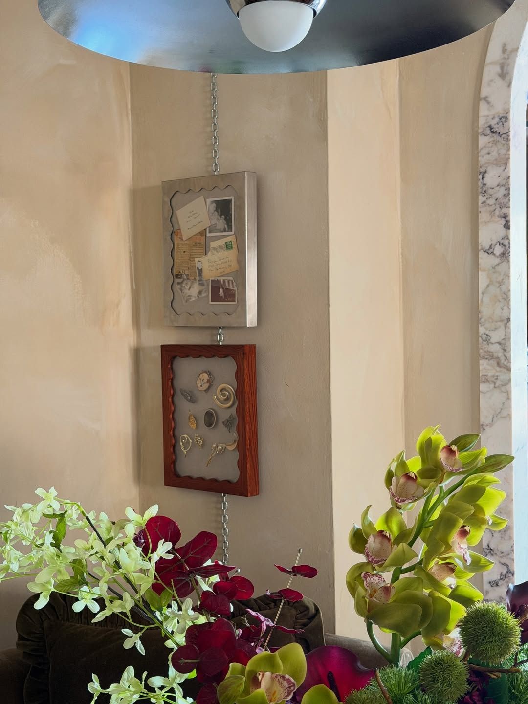 Close-up image of two shadow box window frame hanging on a chain on the wall. The wall is beige limewash and there are plants in the front of the frame.