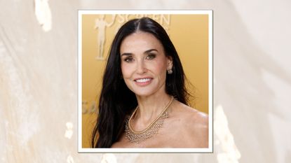 Image of Demi Moore smiling with long straight black hair, tucked one side behind her back, wearing a strapless dress and a diamond-embellished necklace, on a white marble background