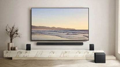 Samsung HW-QS750F soundbar, woofer and surround speakers in muted living room around TV, on marble stand next to plants.