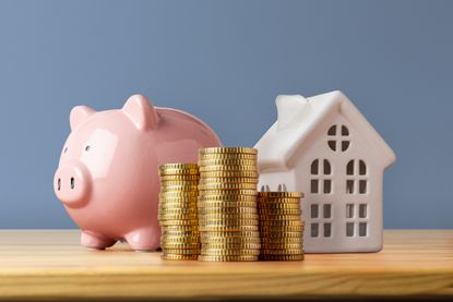 Front view of a wooden table with a piggy bank, a stack of coins, and a house shape against a blue wall.