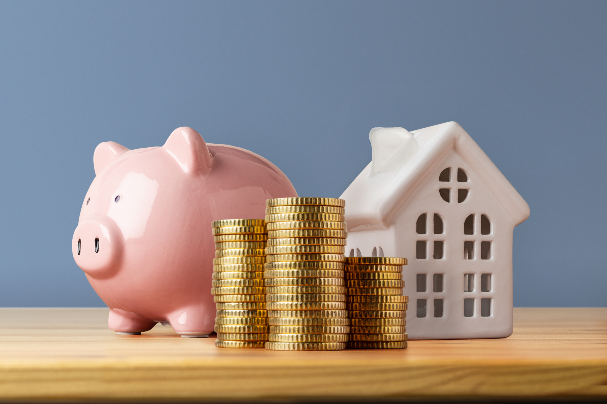 Front view of a wooden table with a piggy bank, a stack of coins, and a house shape against a blue wall.