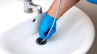 Drain snake being out down white ceramic sink by person wearing blue gloves