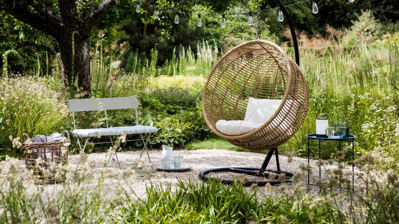 A rattan-effect outdoor hanging egg chair on a gravelled area amongst planting in a mature garden