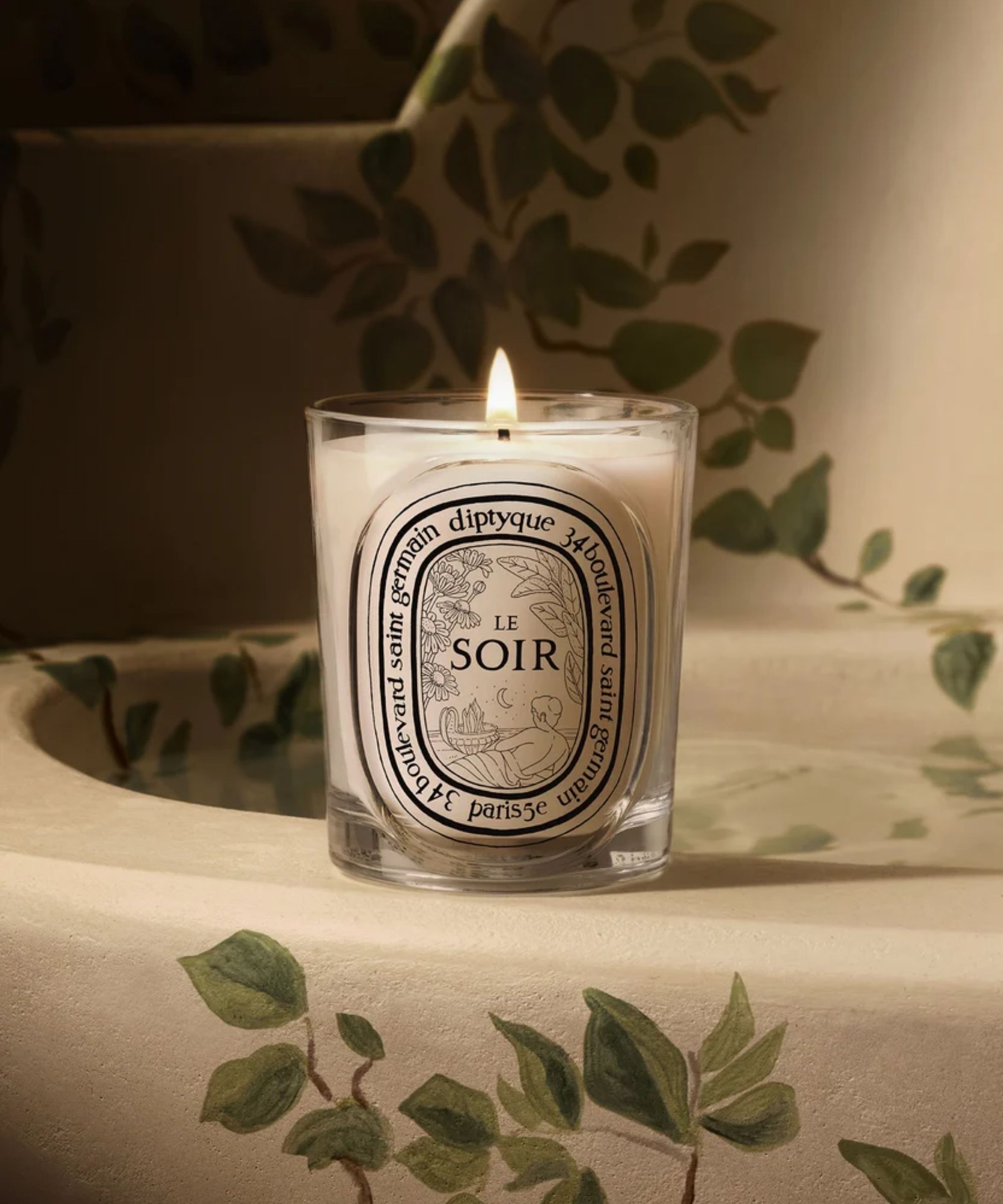 A lit Diptyque Le Soir candle on a white sink in front of a white wall decorated with painted vines