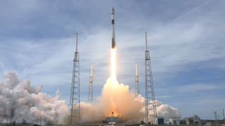 rocket lifting off above plume of fire and smoke. cloudy sky in behind. two launch towers on either side of the rocket 