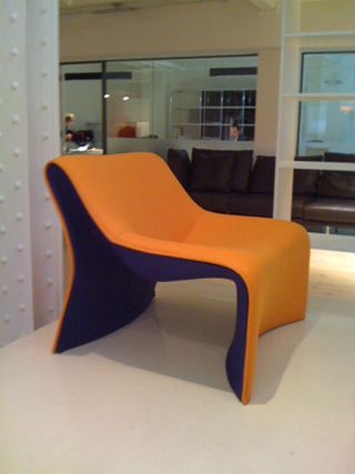 White floor, white metal girders on wall, orange and blue recliner armchair, white frame glass wall, open doorway, blurred brown leather sofa, office, person sat at a desk in the distance