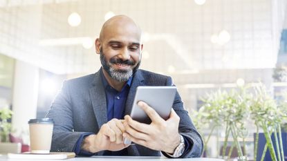 A smiling man looks at his tablet while sitting in a cafe.