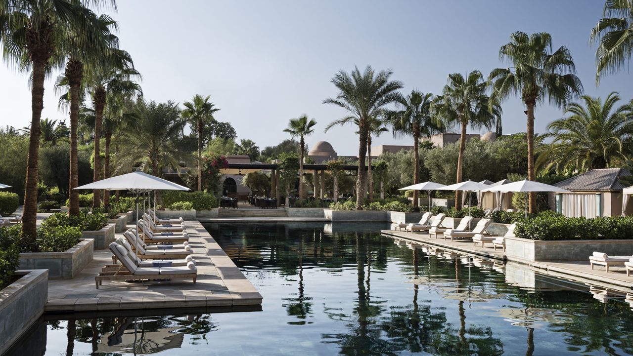 Image of an outdoor pool surrounded by white lounge chairs and umbrella and palm trees.