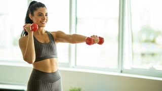 Woman holding a set of dumbbells left arm extended at shoulder height and right arm by head during dumbbell arm workout in studio