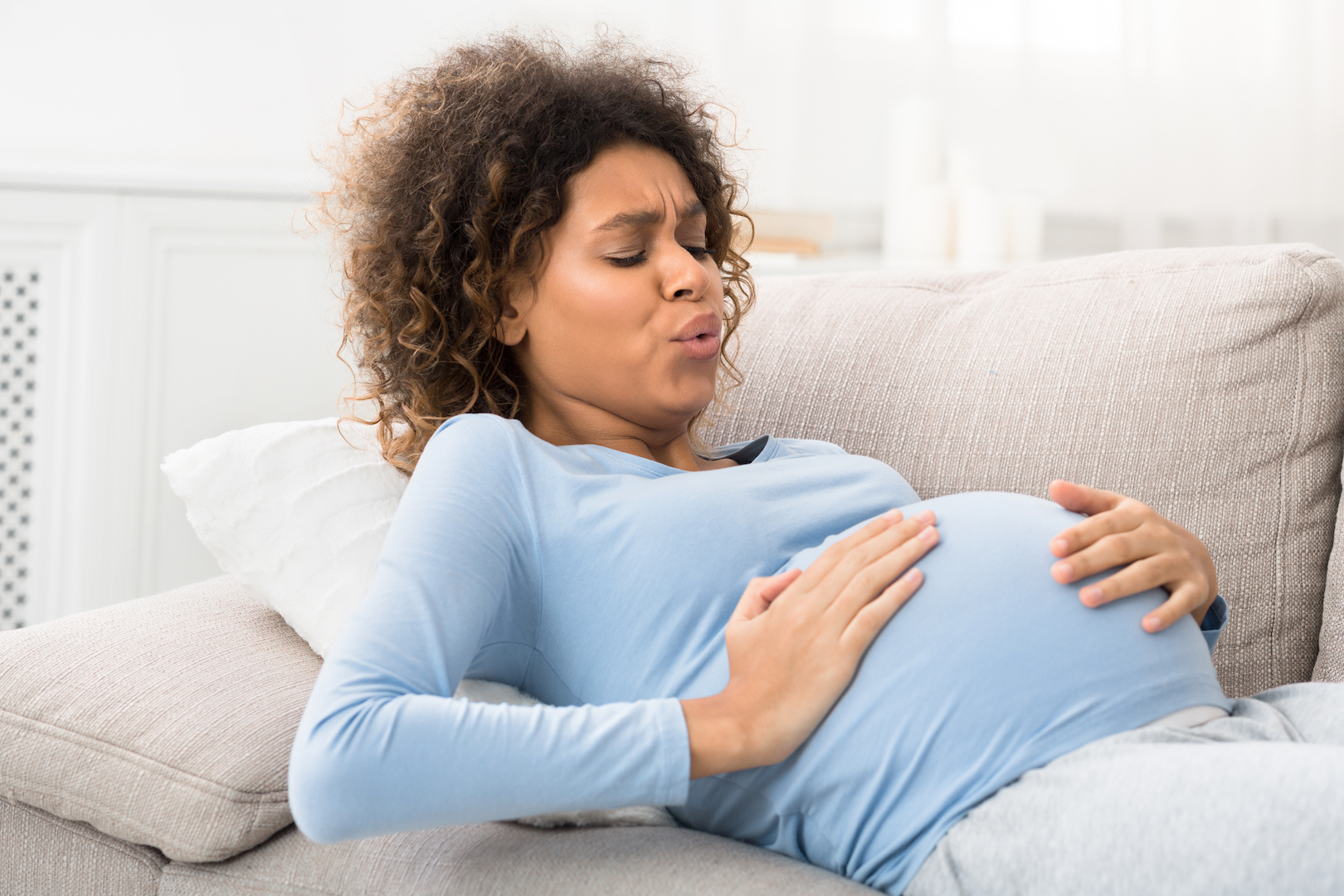 Signs of labor: Pregnant woman winces in pain as she feels a contraction while lying on the couch.