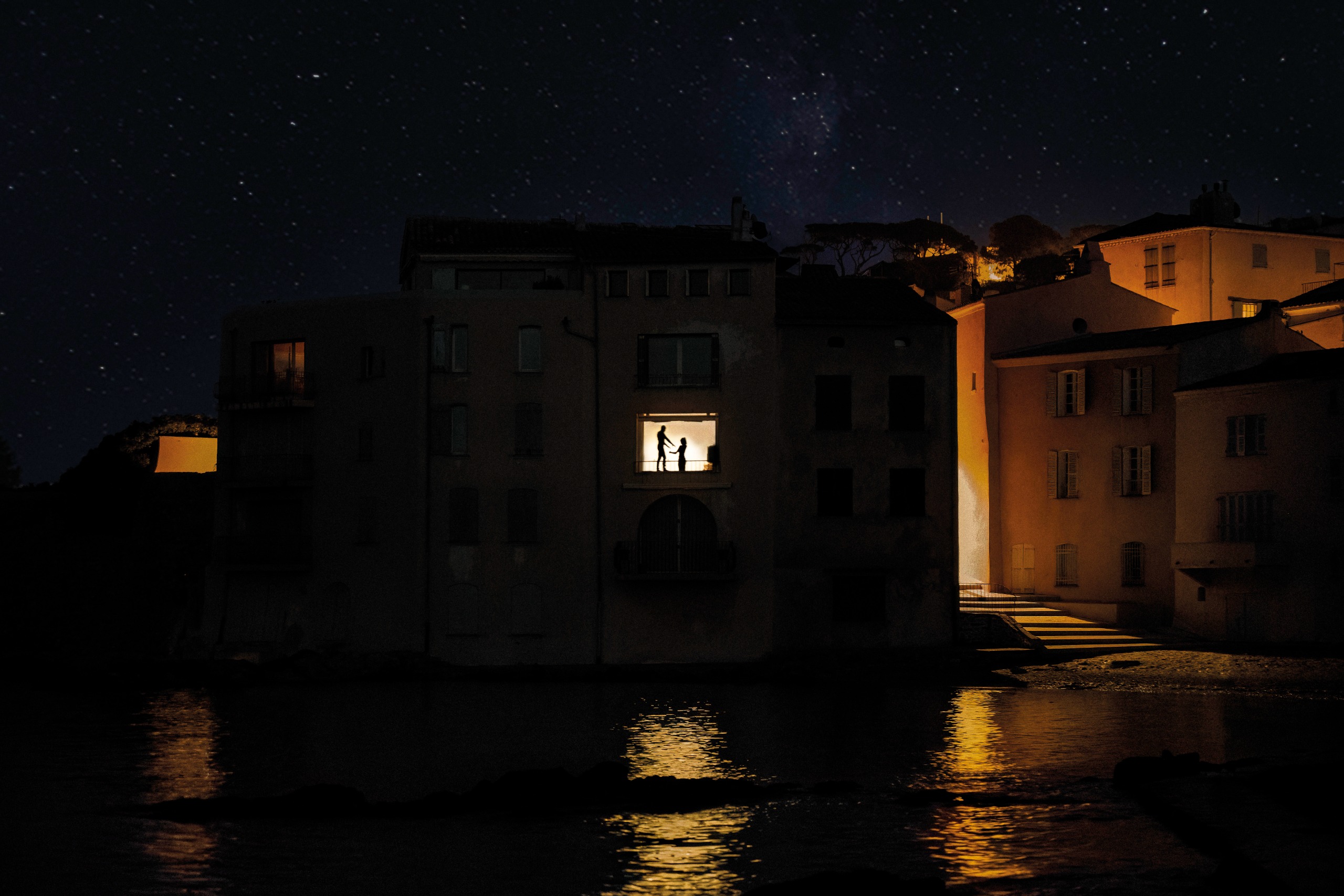 Two figures stand silhouetted in a single lit window of a dark harbourside building, reflected in the still water below beneath a starry sky.