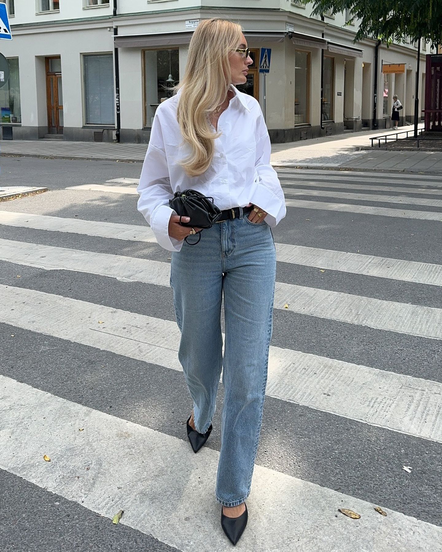 Swedish style influencer Fredrica Eriksson crosses the street wearing a white button-down shirt, small black pouch bag, black belt, straight-leg jeans, and black pointed-toe slingback heels