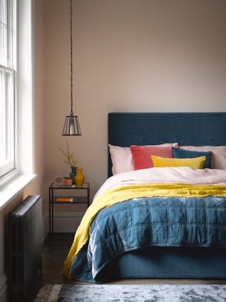 pale pink bedroom with blue upholstered bed, blue quilt, yellow blanket, pink linen bedding and pillows, matching colored cushions, rug, black metal and glass side table, black metal pendant