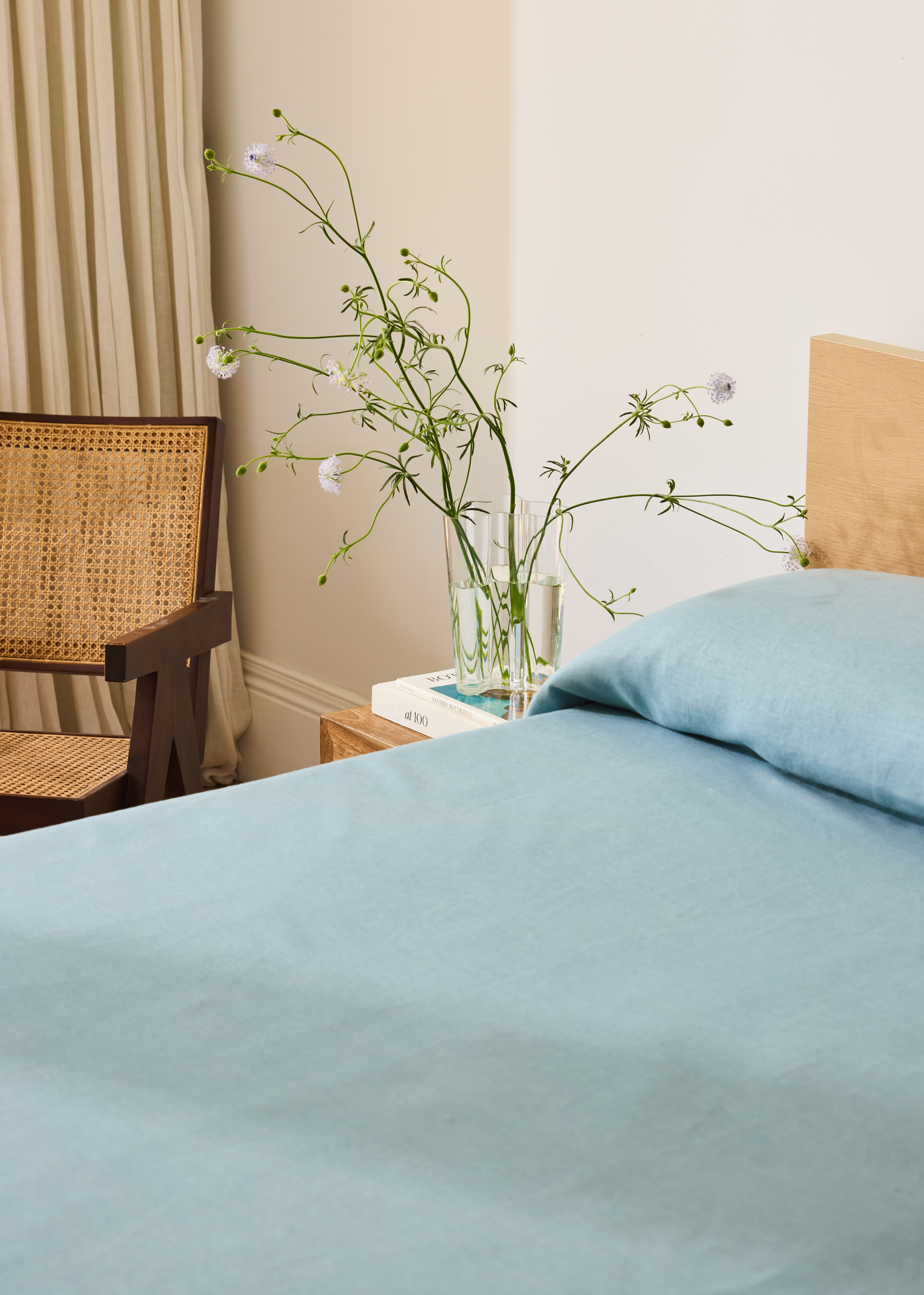 A sky blue bedscape beside a nightstand with a glass vase of tall flowers near a wood and rattan armchair
