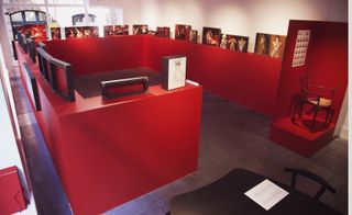 Raised view of display room, slate grey floor, white wall and ceiling with spotlights, windows letting in light far wall, red wall surround display with female erotic art along the top, wooden stool on a red viewing platform with artwork above on a red wall front of shot, black table and chair bottom right