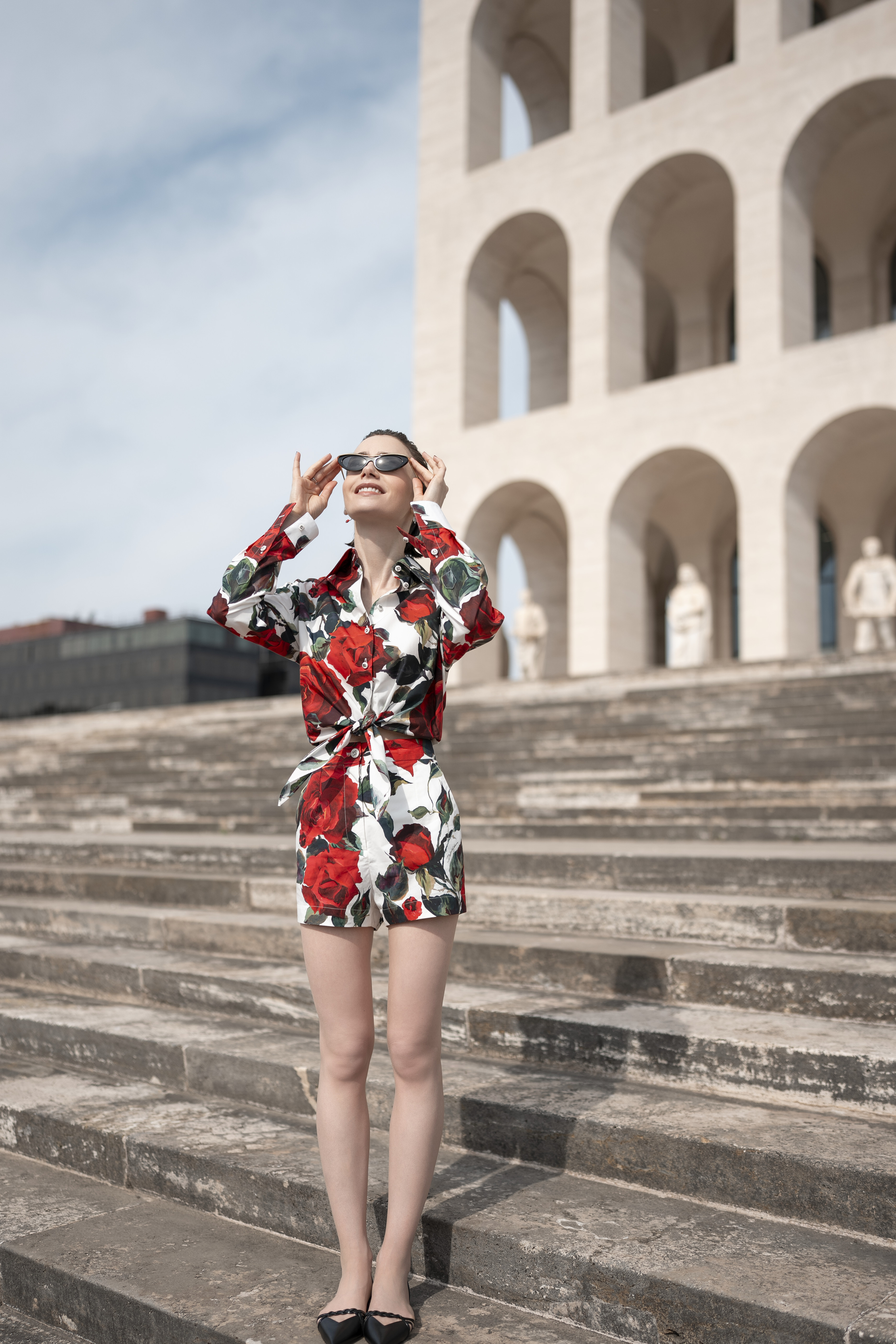 A young woman dressed in a floral print shirt and shorts set in red, white, and green with black flats holds her hands on the sunglasses she wears while facing the sunshine smiling on a staircase in front of an historical, vaulted arches-dotted building.