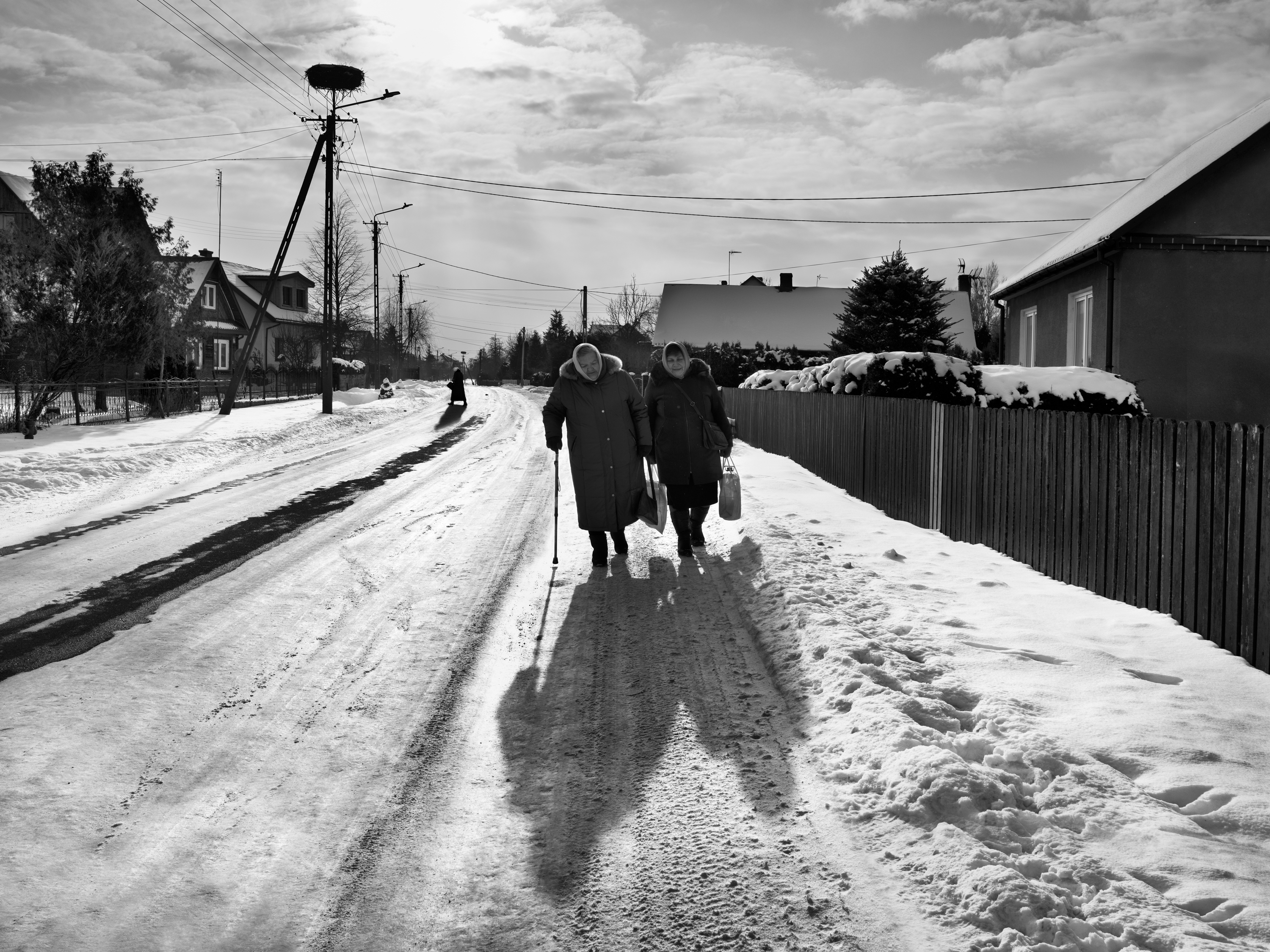 Black and white photograph of two elderly women walking arm in arm along a snow-covered village road, one leaning on a cane, their long shadows stretching ahead of them.