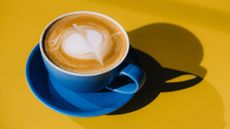 Flat white coffee in blue cup and saucer, sitting on yellow table to represent the benefits of decaf coffee