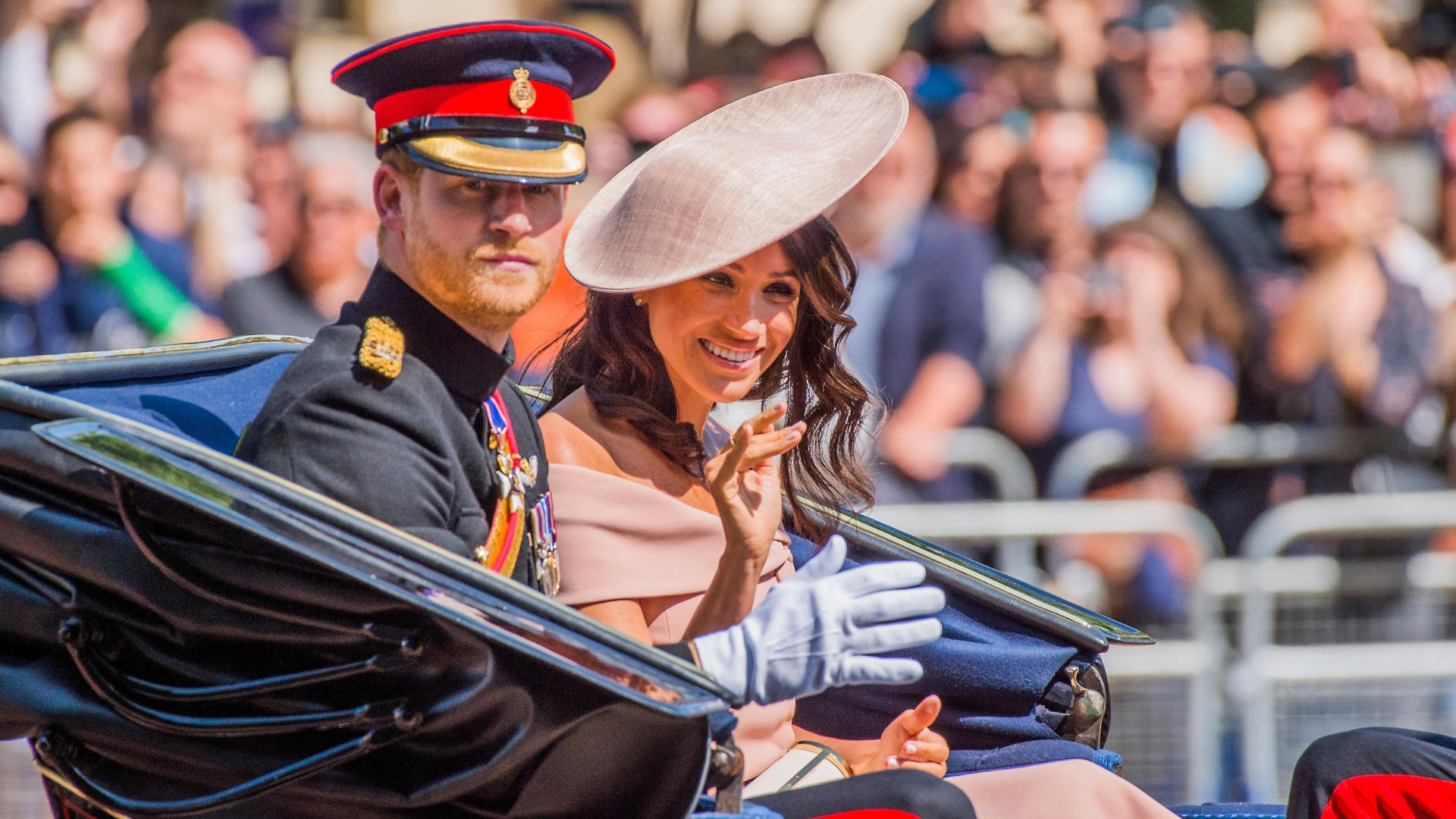 Meghan Sussex wearing a blush pink hat, and Prince Harry wave to fans