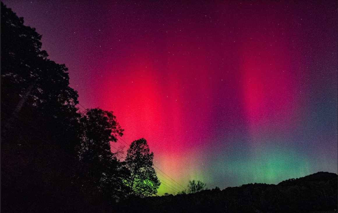 red and green auroras dance above hills and trees in a dark sky