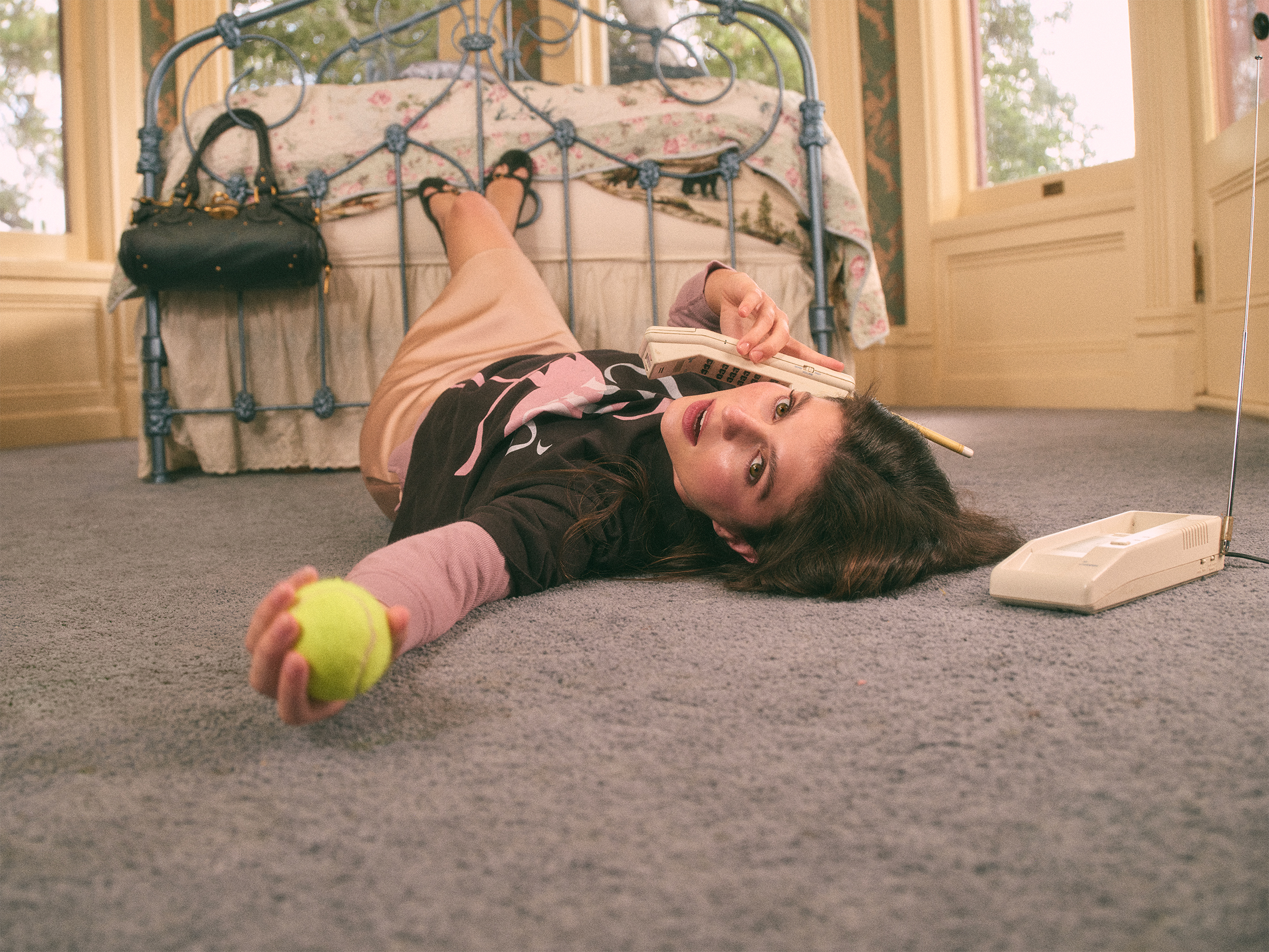 Emily Bader is photographed for Who What Wear's In Focus feature lying on the floor of a bedroom. She is talking on a retro phone with a tennis ball in her other hand, and her feet are perched up on a metal frame bed. She is wearing a Chloe t-shirt with a flamingo on the front and a pink silk skirt.
