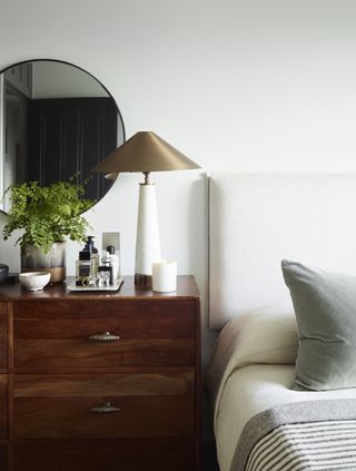 A bedroom with a wooden chest of drawers next to a bed with a lamp on top
