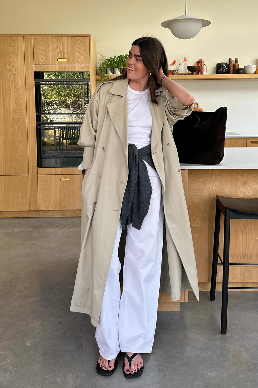 A woman wearing a trench coat, white jeans and black flip flops