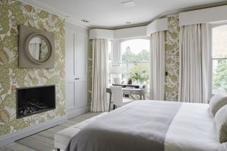 A bedroom with a double bed, a fireplace, patterned wallpaper and a desk in the window