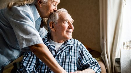 Smiling senior man sitting in an armchair with wife leaning over to kiss him on the head 