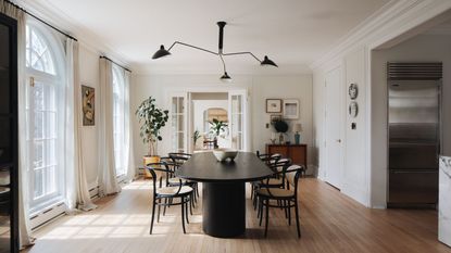Neutral dining room with large dinner table and six chairs, abstract light, white walls and ceiling, two arched doorways and windows with open curtains, and a doorway leading to other room in background. To the right, a large fridge freezer is pictured.