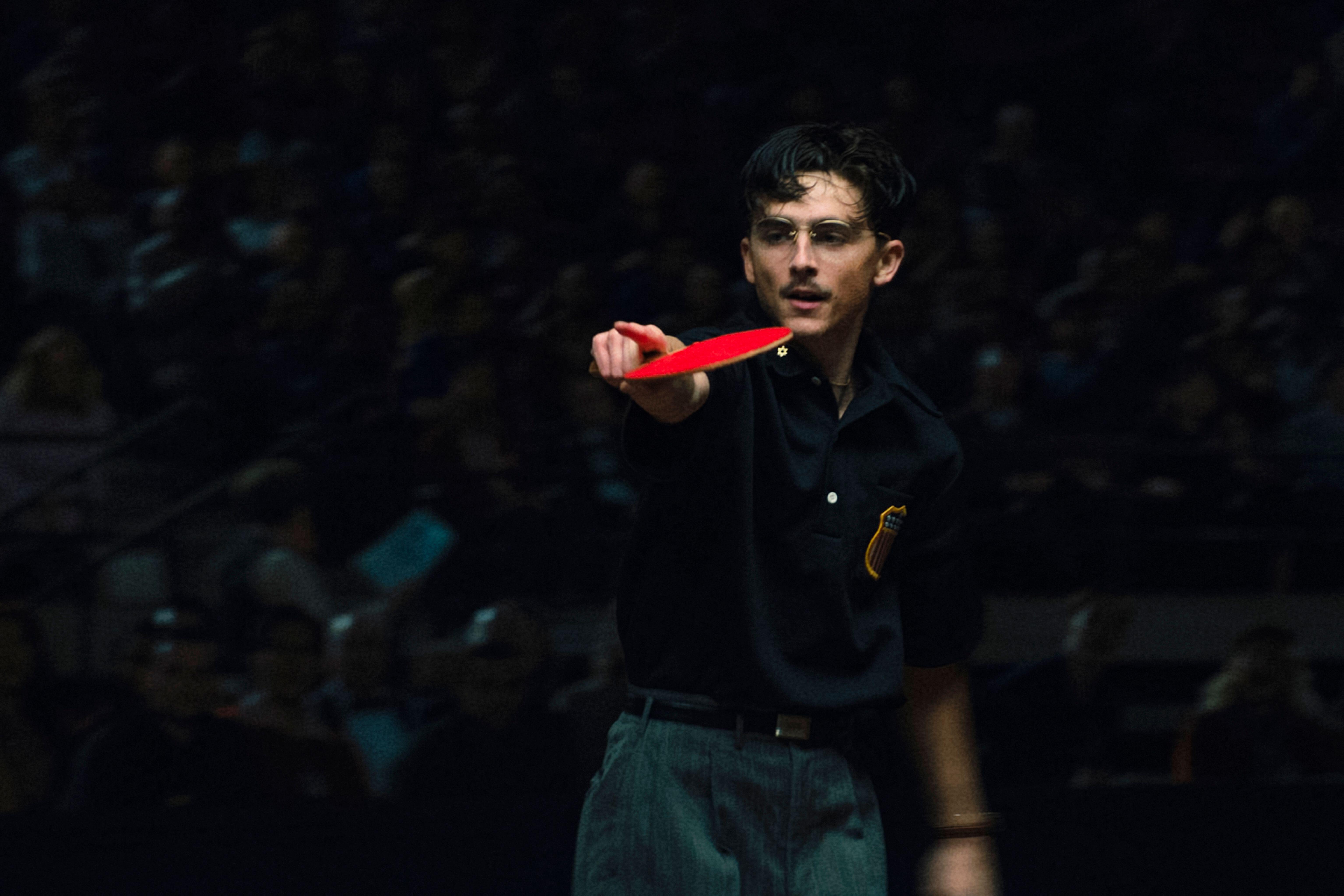 Timothee Chalamet in Marty Supreme holding a racket and pointing at something